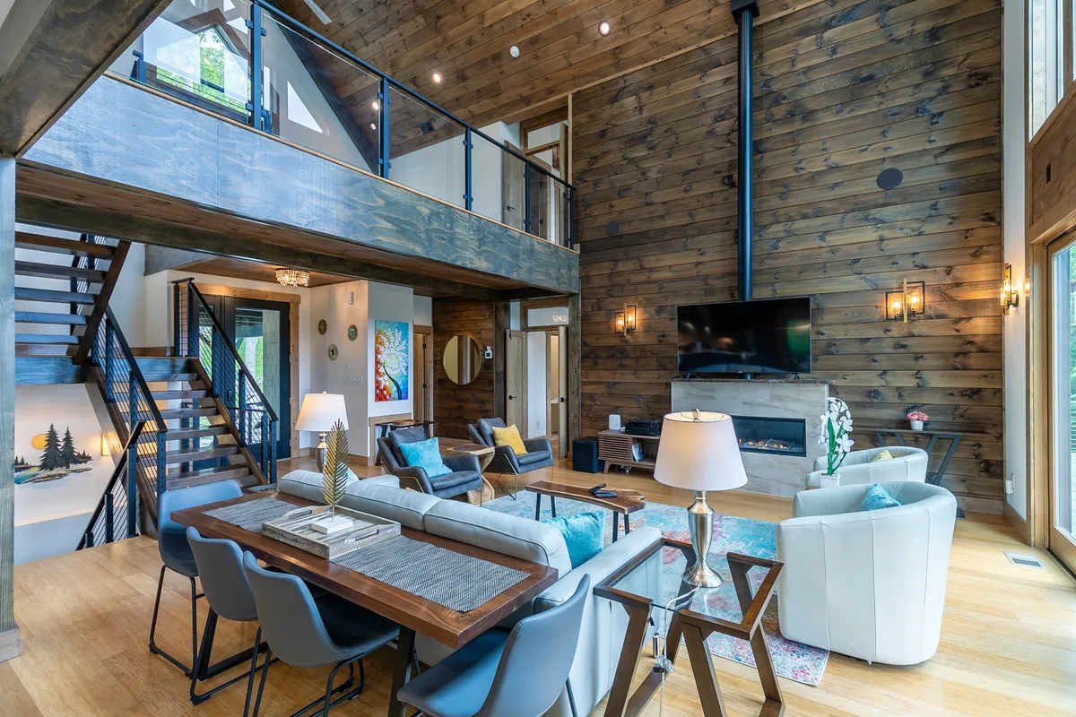 A spacious great room featuring a dramatic wooden beam ceiling, a modern marble-accented fireplace, a generous dining table, and expansive windows that capture sweeping mountain views. The open-concept design blends living and dining areas, with soft natural light enhancing the warm, inviting atmosphere.