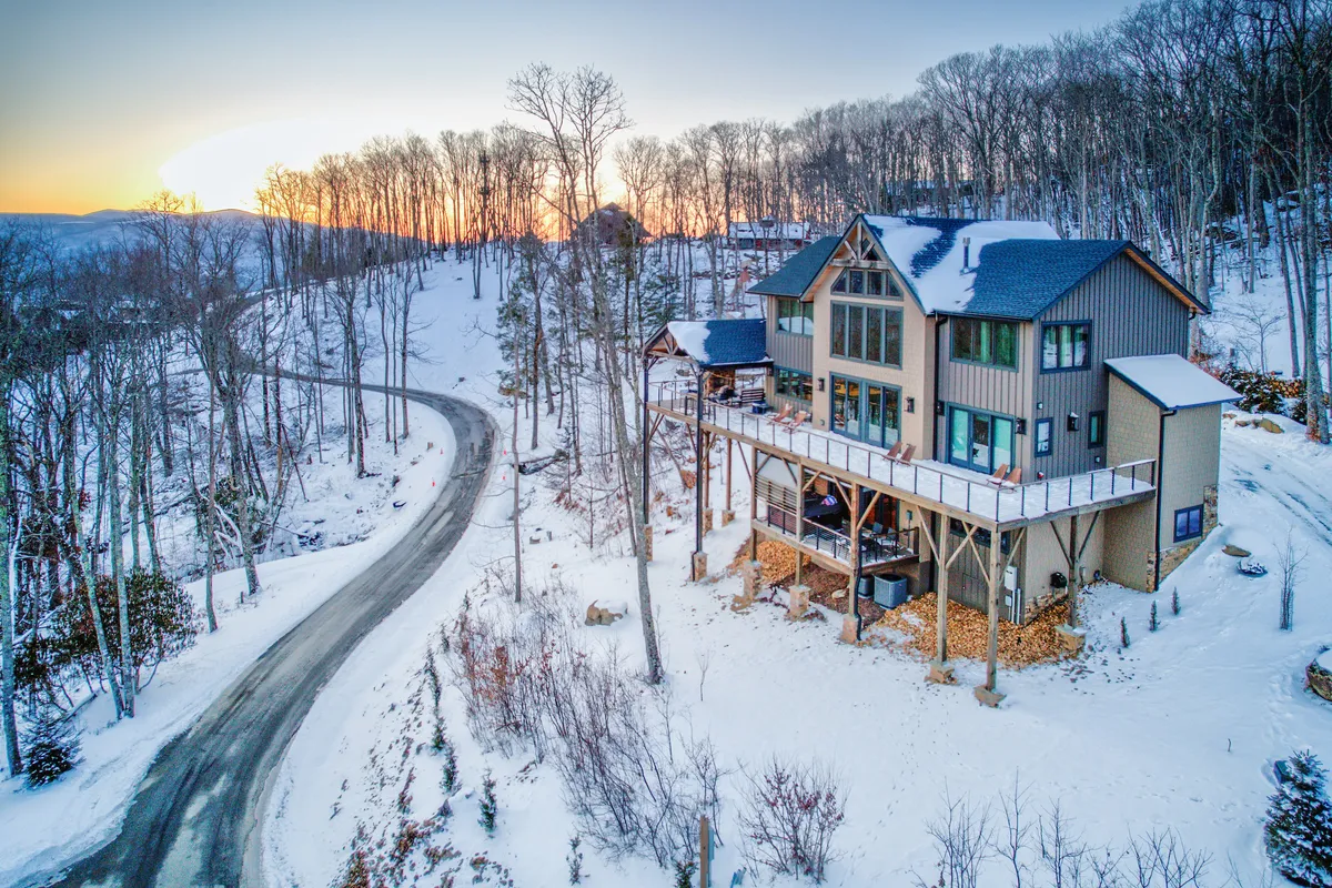 A tranquil winter scene showing the house framed by snow-covered trees and a winding road. The landscape exudes calmness, with opportunities for outdoor winter activities like skiing and snowshoeing, as well as relaxing on the porch while soaking in the snowy ambiance.