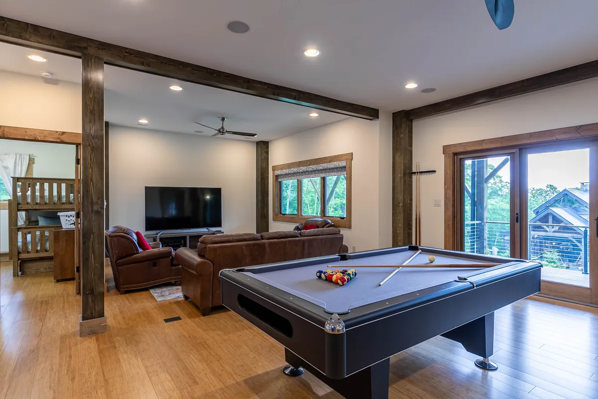A vibrant game and media room featuring a pool table, a plush sofa, a large TV mounted on the wall, and sliding glass doors that flood the space with natural light. The layout encourages both friendly competition at the pool table and relaxed movie nights on the couch, with a glimpse of the outdoors through the sliding doors.