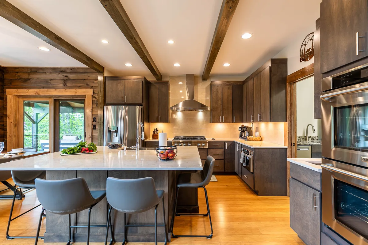 This image presents a spacious, modern kitchen featuring a large kitchen island, stainless steel appliances, prominent wooden beams overhead, and an open-concept layout. The kitchen is designed for both culinary adventures and socializing, highlighted by the generous counter space and luxury appliances.