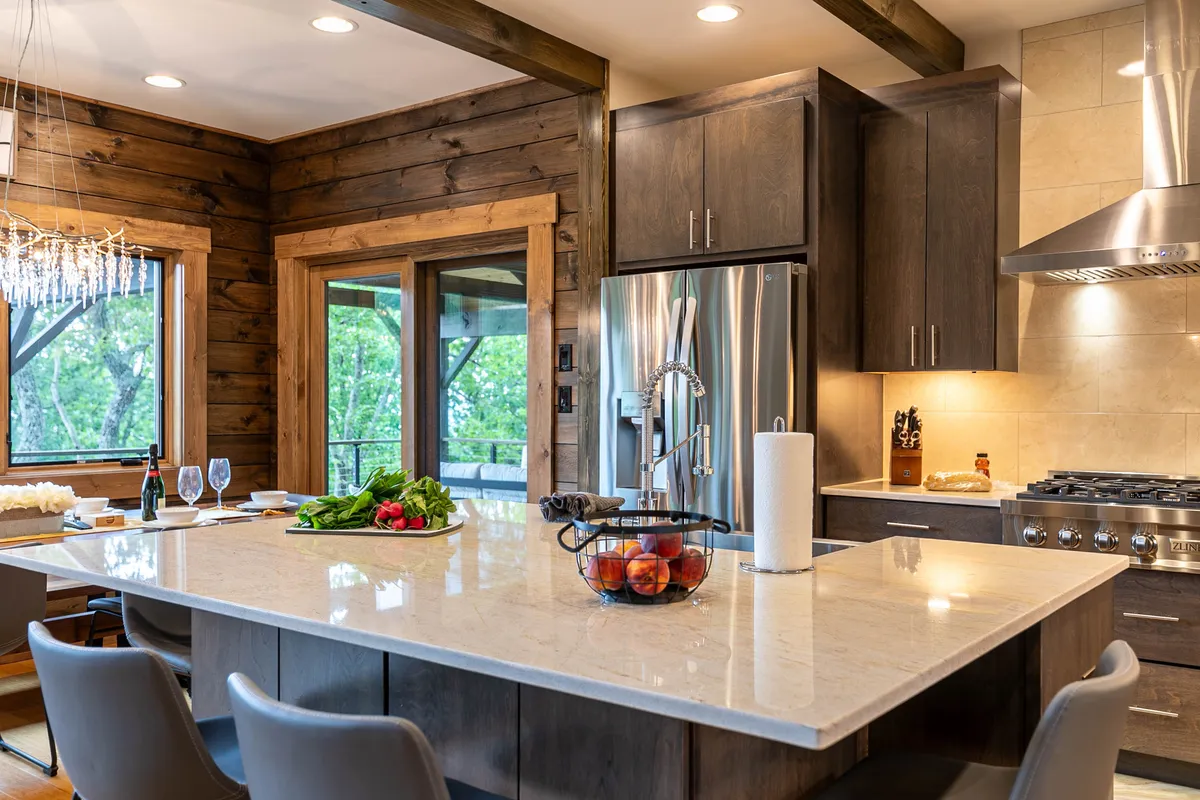 A welcoming and contemporary kitchen scene, this image features a large kitchen island, stainless steel appliances, a sparkling chandelier, and windows that frame views of lush greenery. Fresh produce displayed on the counter adds to the sense of culinary abundance and hospitality.