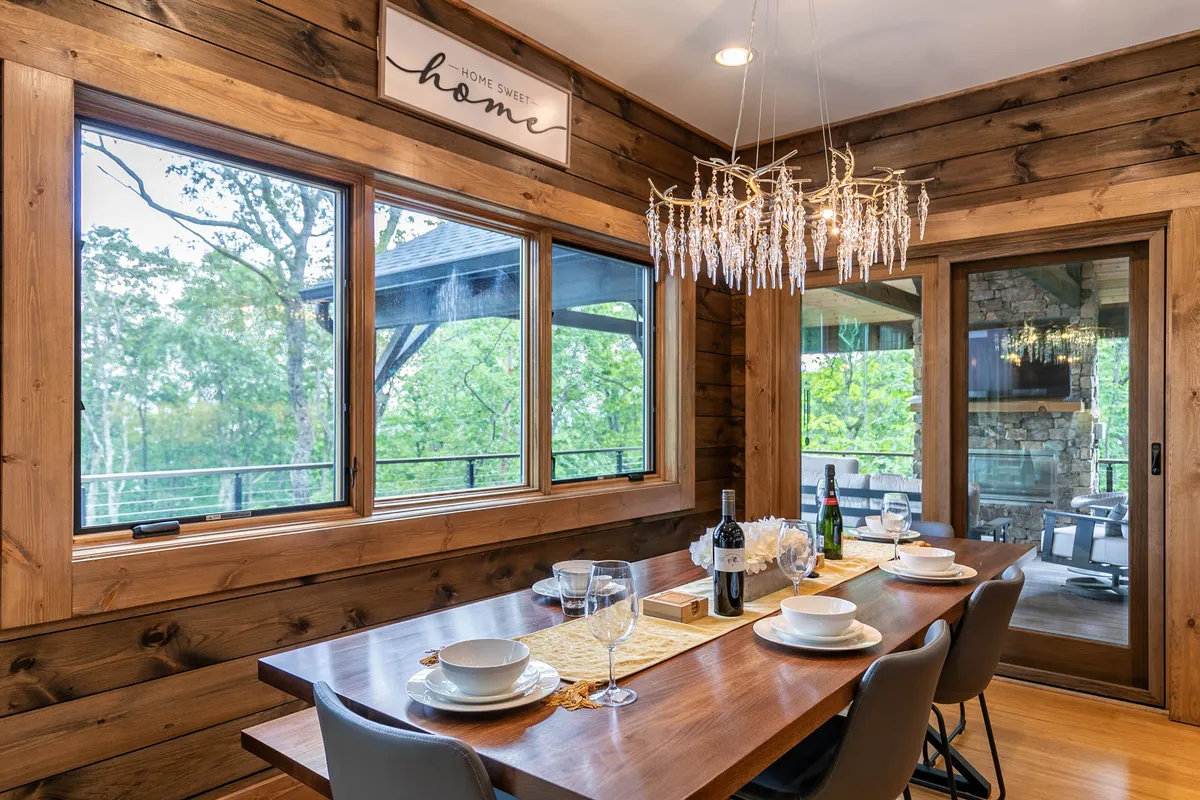 This cozy dining area centers on a large wooden dining table beneath an elegant chandelier, with windows offering scenic views. The table is set for a meal, inviting gatherings and shared experiences.