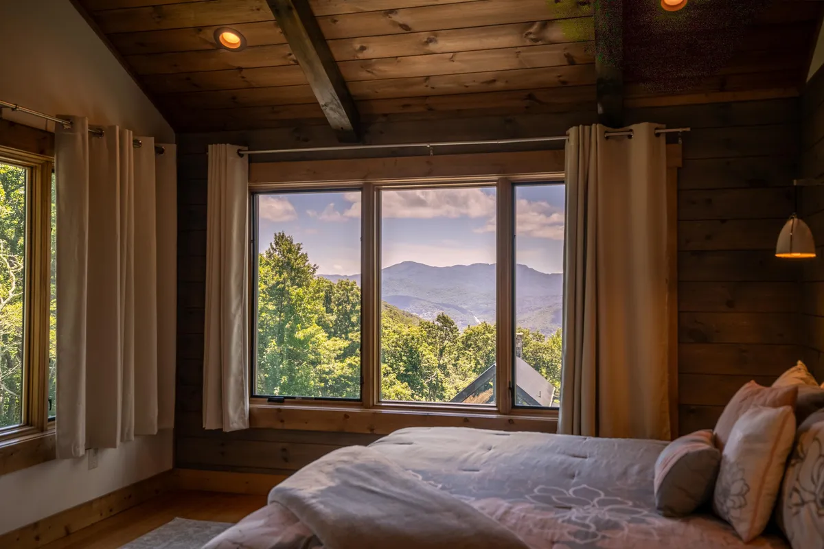 A spacious bedroom with a large bed adorned with decorative pillows, wooden ceiling beams, and floor-to-ceiling windows that display breathtaking mountain vistas. The soft natural light and luxurious linens create an inviting environment for relaxation.