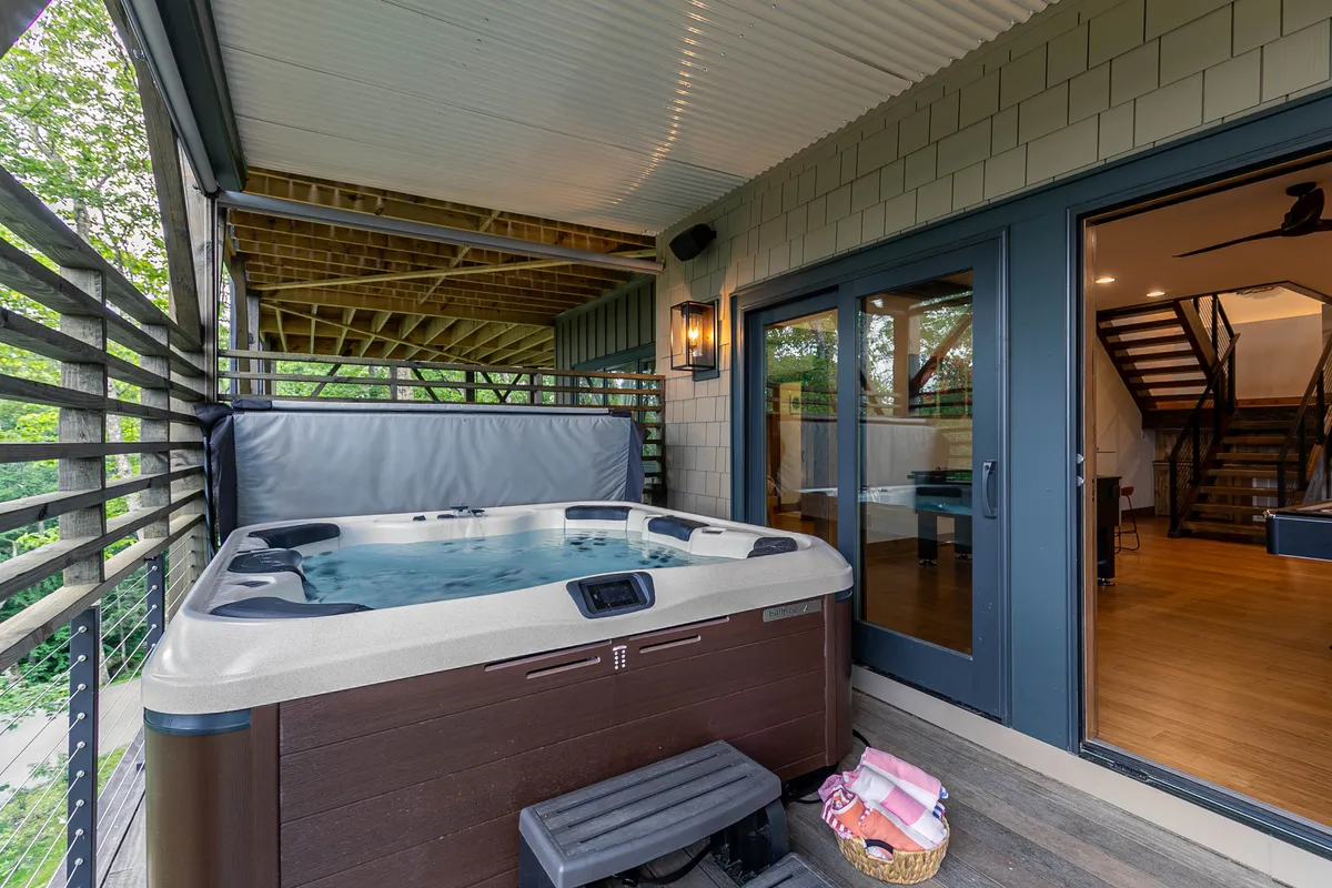 A spacious wooden deck features a bubbling hot tub with adjacent seating, all set against a backdrop of lush greenery and large windows that hint at the comfort inside. The setup suggests an ideal spot for group relaxation after a day of vineyard tours or outdoor fun.